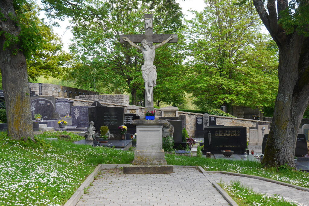 Hinter einem mächtigen Kreuz mit Corpus Christi aus grauem Sandstein ziehen sich ausschließlich dunkle Grabmäler einen leichten Hang nach oben.