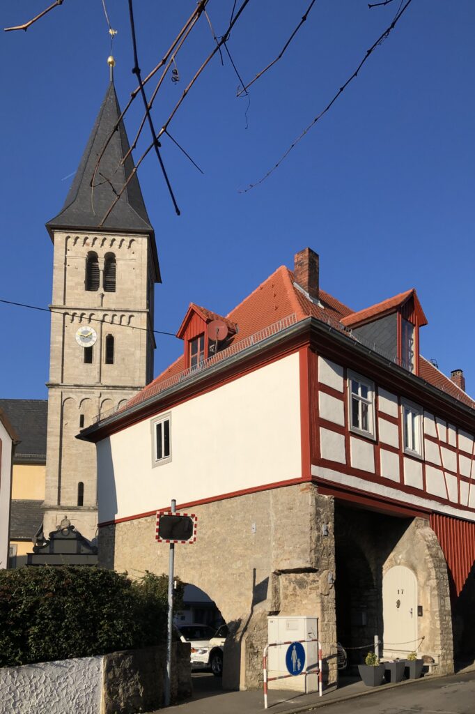 Kirchturm mit Stilelementen aus Romanik und Gotik, davor ein Gebäude mit rotem Fachwerk samt Tordurchfahrt.