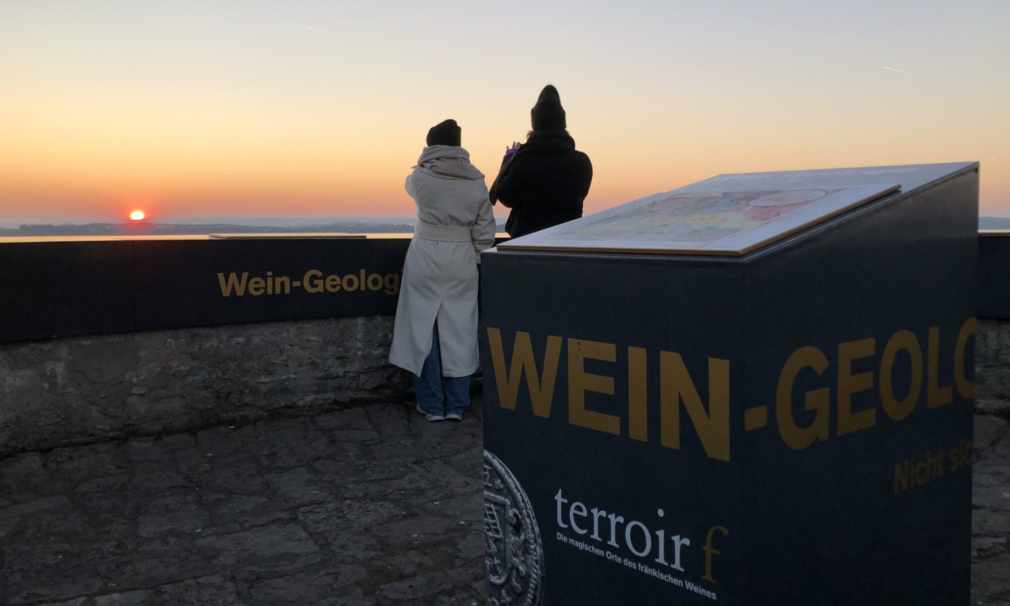 Zwei warm gekleidete Personen schauen zur untergehenden Sonne am Horizont. Ihr Aussichtspunkt ist der terroir f auf dem Randersackerer Sonnenstuhl.