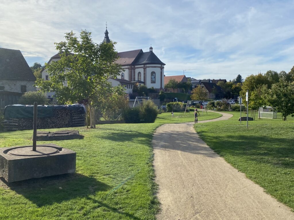 Die Talaue, wo sich der Dorfweiher befand, ist begrünt und mit Spielgeräten für Jung und Alt ausgestattet. Oberhalb steht die Pfarrkirche St. Margaretha.