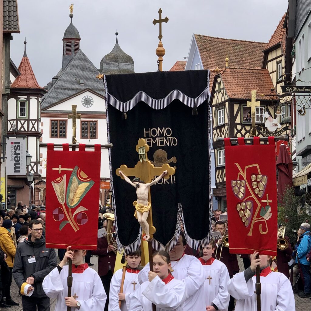 Ministranten führen eine Prozession an. Einer trägt eine schwarze Fahne mit der Aufschrift "Homo Memento Mori".