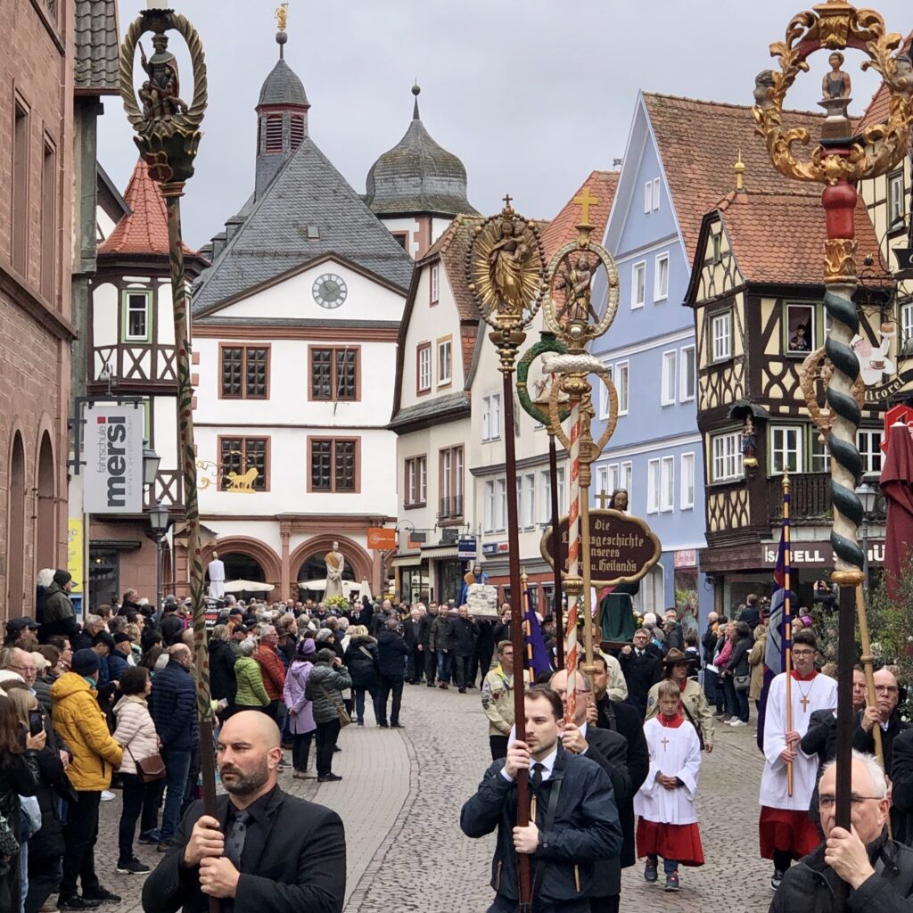 Dunkel gekleidete Männer tragen geschnitzte Symbole für Handwerksberufe. Diese sind auf Stangen gesteckt, damit sie hoch aufragen und gut sichtbar sind.
