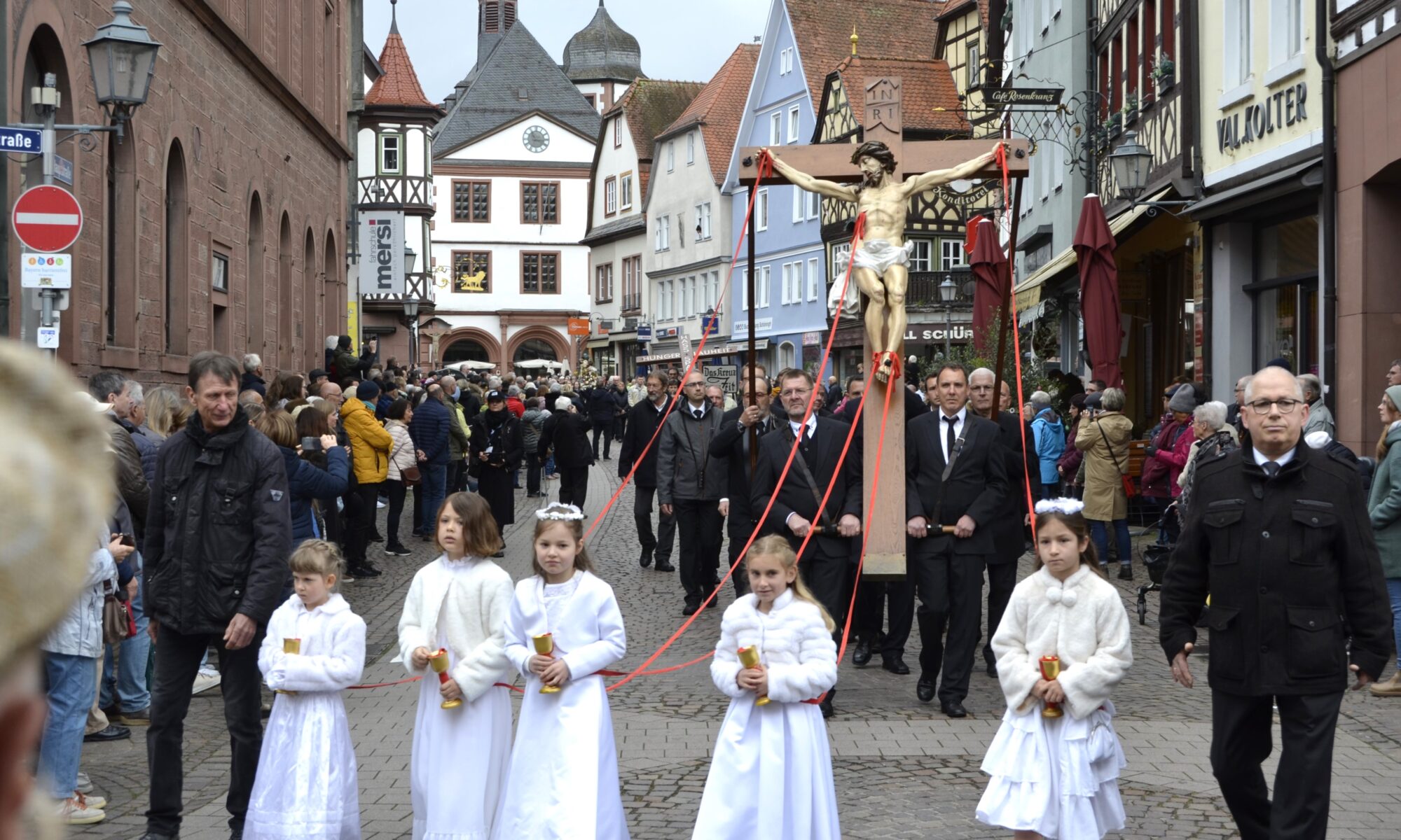 Männer tragen eine lebengroße Jesusfigur am Kreuz durch eine menschengesäumte Straße. Vorneweg gehen Mädchen in weißen Kleidern.